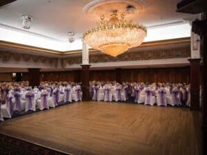 A grand ballroom is set up for a formal event. White covered chairs with purple ribbon sashes are arranged in rows around a large, polished wooden dance floor. A massive, intricate chandelier hangs from the ceiling, illuminating the space. Elegant gold drapery decorates the walls and columns, making it an ideal wedding venue.