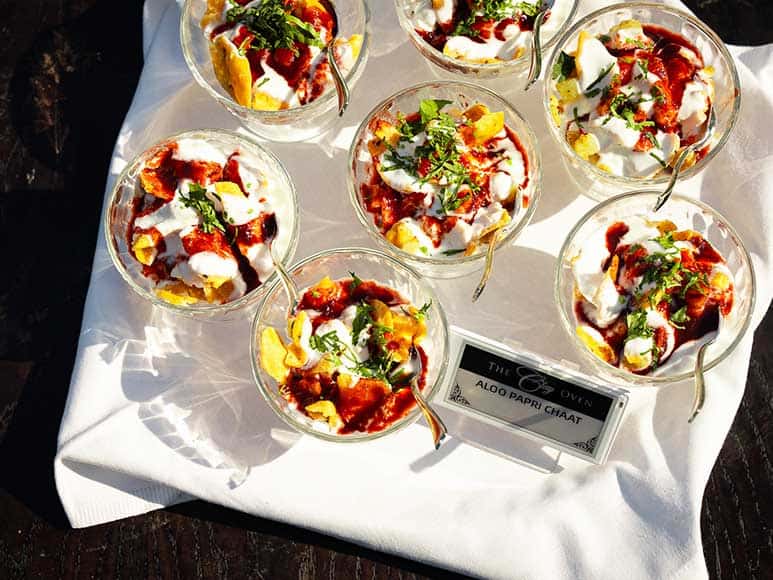 Close-up of several glass bowls filled with Aloo Papri Chaat, perfect for an event catering setup, placed on a white cloth. The chaat is topped with yogurt, tamarind chutney, chopped coriander, and sev. A small sign next to the bowls reads “Aloo Papri Chaat.” Silver spoons are inserted into each bowl.