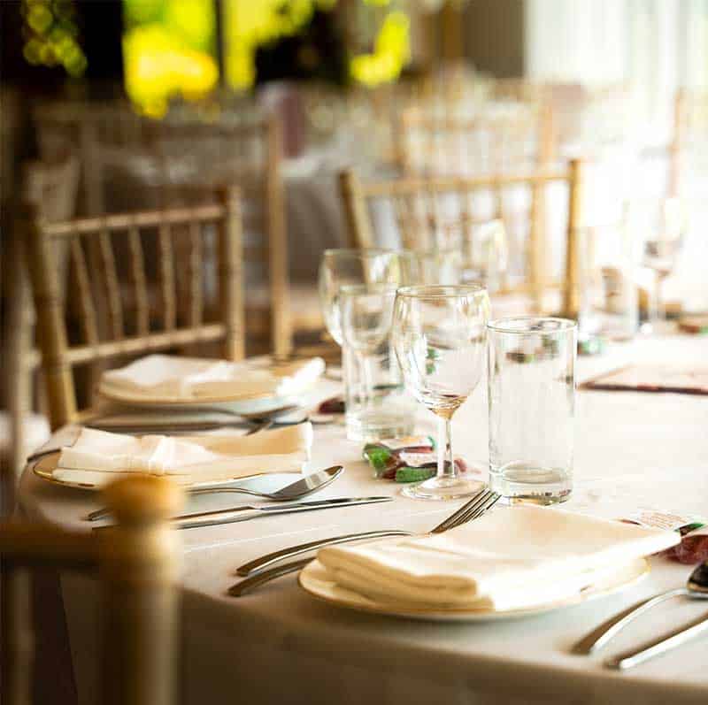 A round dining table set for an elegant event. The table is covered with a white tablecloth, and place settings include folded white napkins, cutlery, wine glasses, water glasses, and small plates. The background shows more similarly set tables by Asian wedding caterers and blurred, soft lighting.