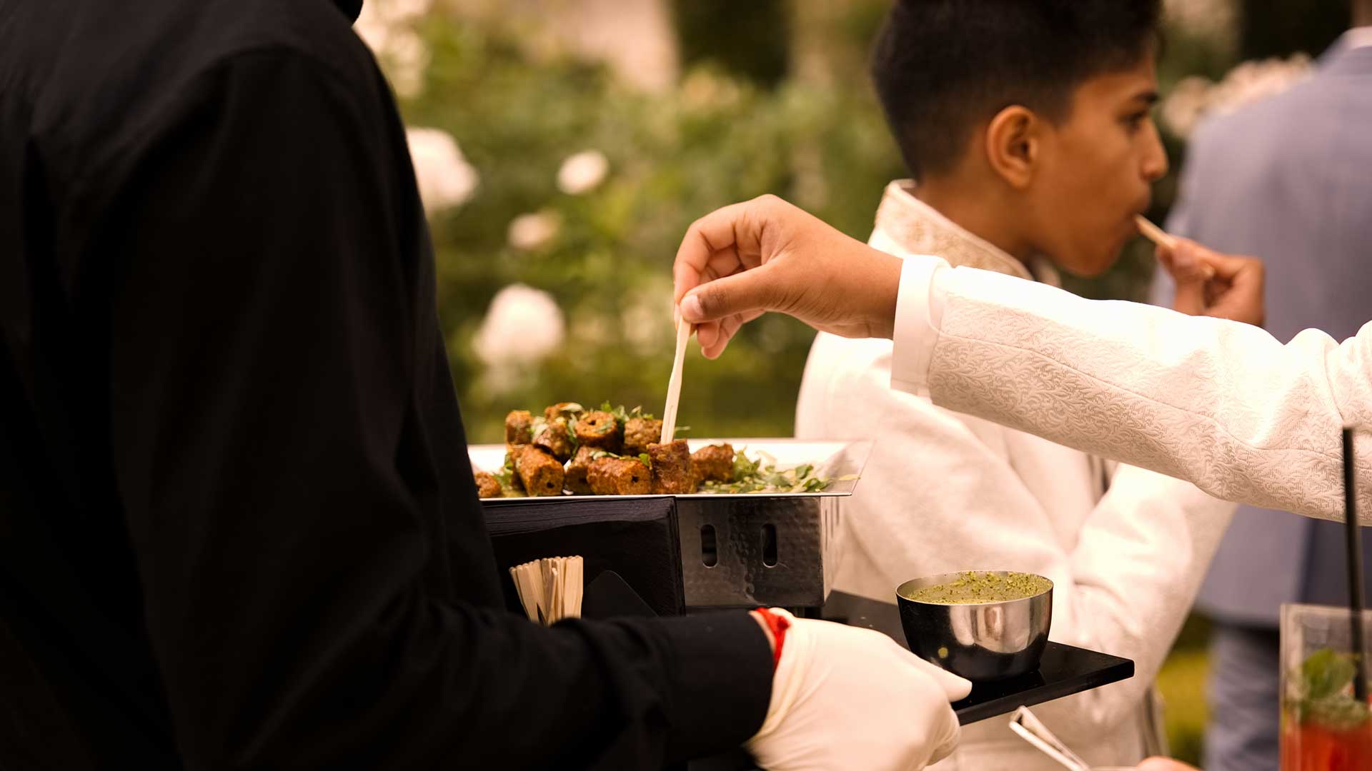 A person in a white jacket reaches for skewered hors d'oeuvres from a tray held by a server in black. Another person in the background, wearing a white outfit, is seen with food, against an outdoor garden setting with green foliage and white flowers.