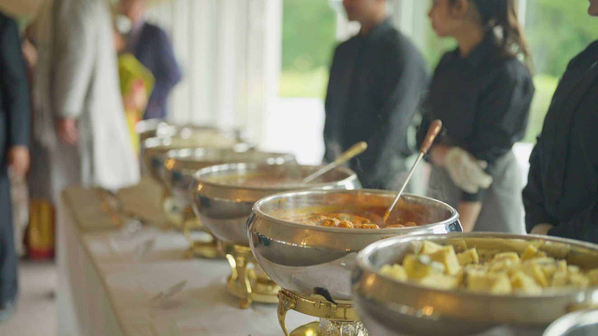 A buffet table is laid out with various dishes in large silver chafing dishes. Two servers in black uniforms and white gloves stand behind the table, ready to assist. In the background, partially visible guests are speaking with one another. The scene appears to be at a catered event.