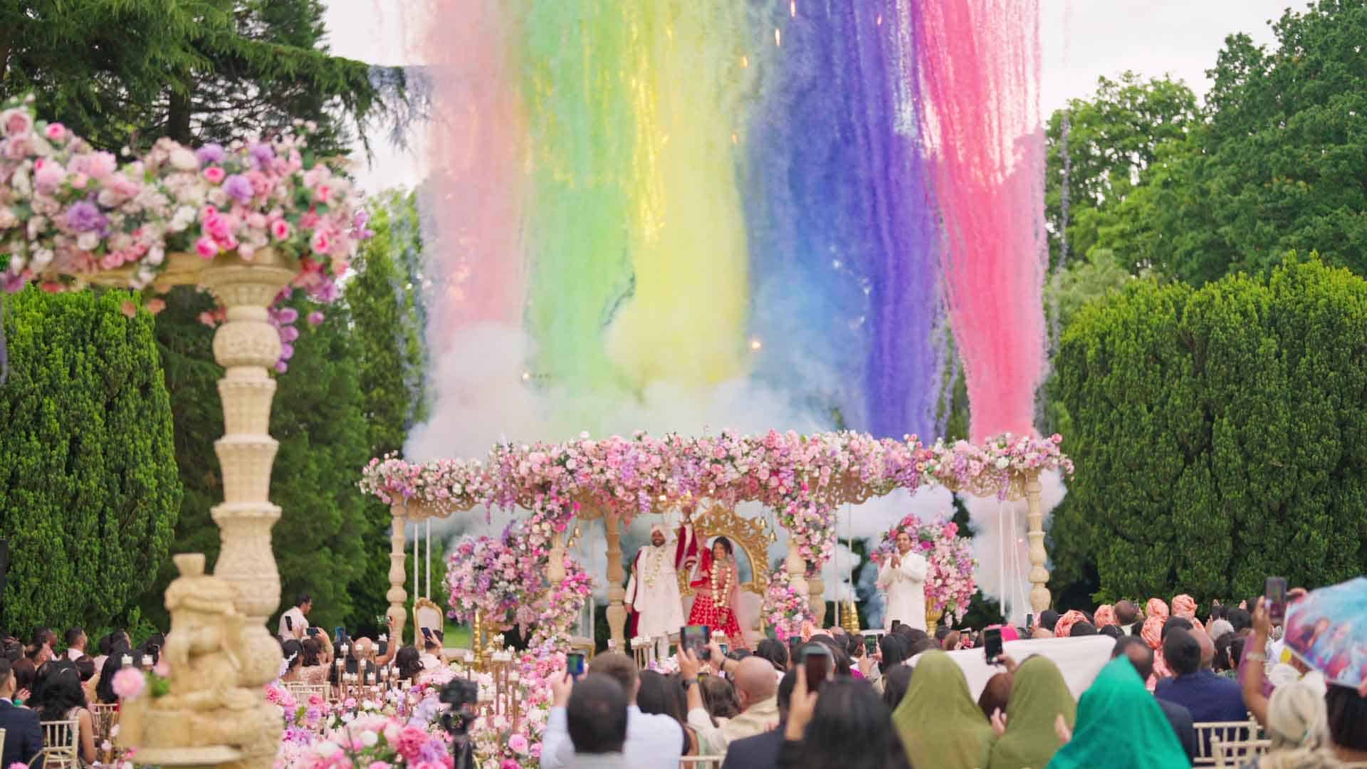 A vibrant outdoor wedding ceremony is underway with guests seated amidst lush greenery. The couple stands under a floral archway adorned with pink flowers. Above them, a canopy of colorful powder in shades of blue, pink, green, and yellow is dramatically cascading down.