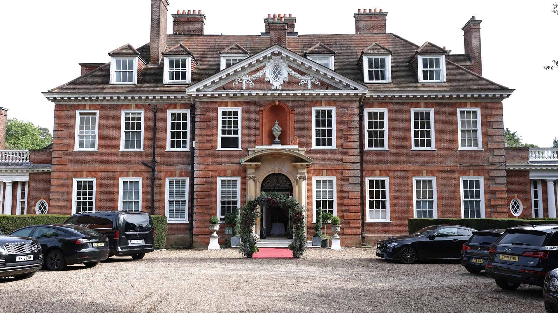 A large, elegant red-brick mansion with a symmetrical design, featuring numerous tall windows and a central entrance adorned with greenery. A gravel driveway in front has several parked luxury cars. The building is surrounded by well-kept hedges and trees.