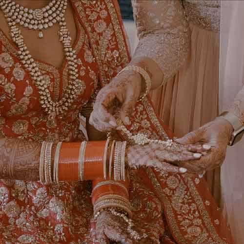 A close-up of an Indian bride in a red and gold embroidered lehenga at the wedding venue. She is adorned with traditional jewelry and red and gold bangles. Her hands, decorated with intricate henna designs, are being assisted by another person, also in traditional attire, who is adjusting her bracelet.