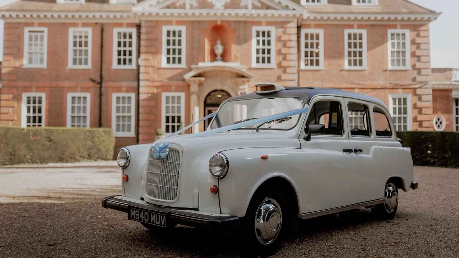 A classic white taxi with a vintage license plate "1940 MUW" is parked on a gravel driveway in front of a large, historic brick mansion. The mansion has numerous windows, decorative white trim, and a grand entrance with an arched doorway.