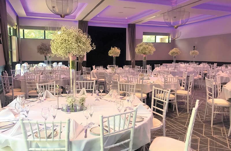 A banquet hall at Denham Grove with round tables set for an event. Each table has white linens, glassware, napkins, and a tall centerpiece of baby's breath flowers. White chairs surround the tables, lit by soft purple ceiling lights by The Clay Oven.
