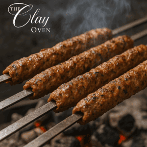 Four skewers of seasoned minced meat kebabs grill over hot charcoal, with smoke rising. The kebabs’ texture reveals herbs and spices. “The Clay Oven” in elegant white script hints at a tandoor event experience. The background is blurred.