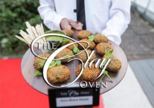 A server in a white shirt and black tie holds a round wooden platter with green-flecked kebabs, fresh greens, and bamboo picks—perfect for wedding catering. A "The Clay Oven" sign is visible above a red surface below the platter.