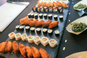 A slate platter displays assorted sushi rolls and inari sushi in neat rows, perfect for event catering or elegant restaurant tables. The rolls vary in filling and topping, with small dishes of seaweed salad on the sides against a dark table background.