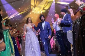 A bride in a sparkling white gown and a groom in a navy suit walk hand-in-hand down an aisle, smiling, surrounded by cheering guests. Multicolored confetti falls as vibrant lighting and joyful expressions set the scene for unforgettable wedding catering.