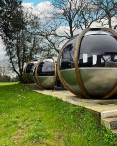 Three modern Pods by Clay Oven with reflective windows and wooden frames sit on a stone patio, surrounded by green grass. Tall trees and a partly cloudy sky create the perfect Romantic Outdoor Restaurant setting for a Date Night in Denham.