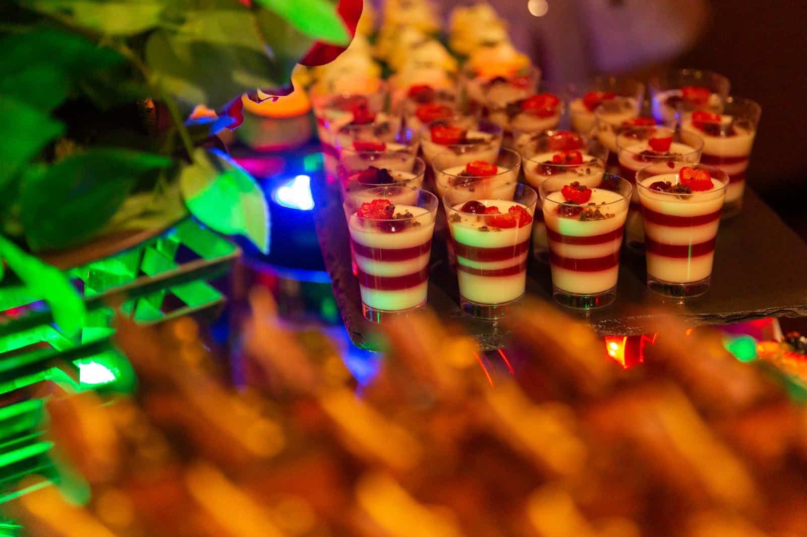 Rows of small dessert glasses filled with layered white and red mousse are arranged neatly on a reflective surface. Each dessert is topped with red fruit and chocolate pieces. Bright, colorful lights and green leaves decorate the background.