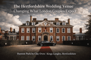 A grand Georgian-style mansion with tall windows and columns stands under a dramatic sky. A black luxury car and red carpet welcome London couples to Hunton Park, a notable Hertfordshire wedding venue perfect for any special occasion.